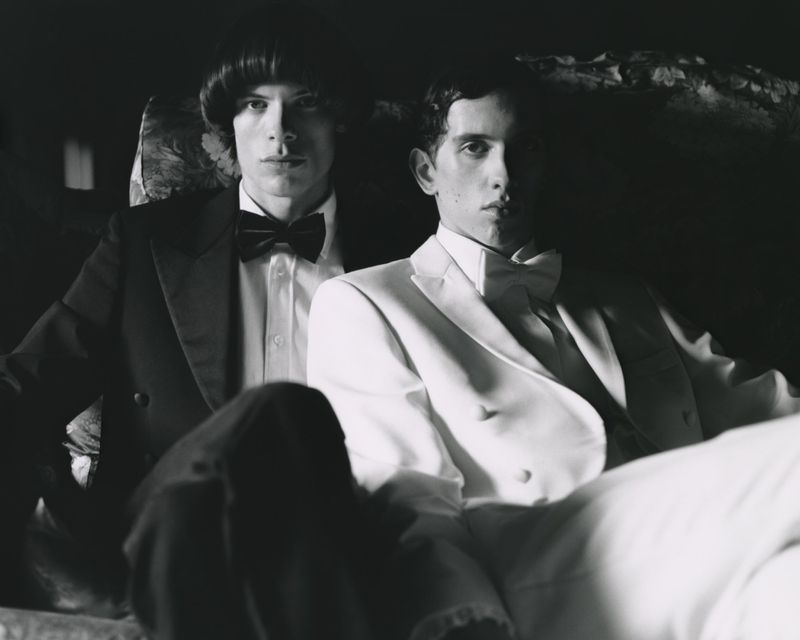 Monochrome portrait of two adult men in formal wear. One man in a black tuxedo with bow tie, the other in a white tuxedo.