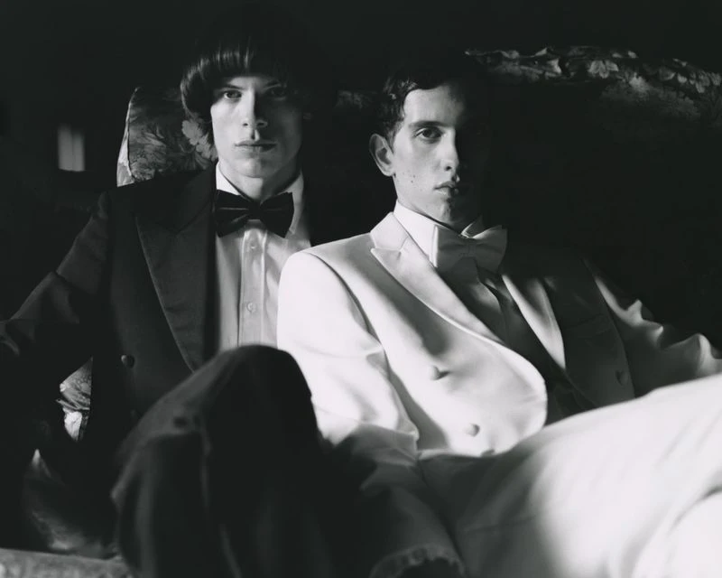Monochrome portrait of two adult men in formal wear. One man in a black tuxedo with bow tie, the other in a white tuxedo.