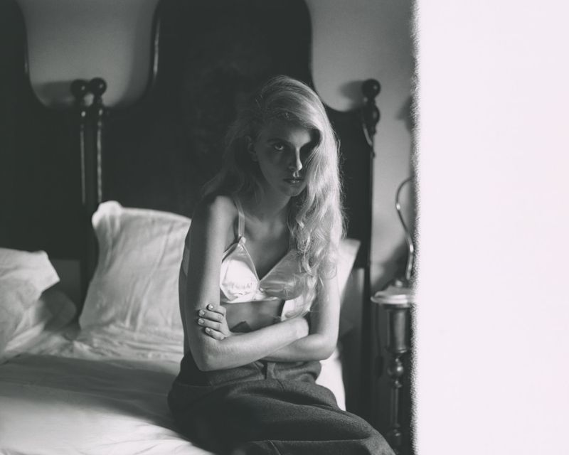 Monochrome photograph of a blonde woman sitting on a bed with folded arms, wearing a light satin bra and dark trousers.
