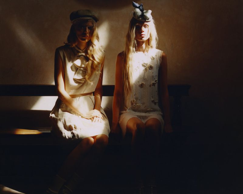 Two women in cream sleeveless dresses and vintage hats sit on a wooden bench illuminated by a sharp beam of light.