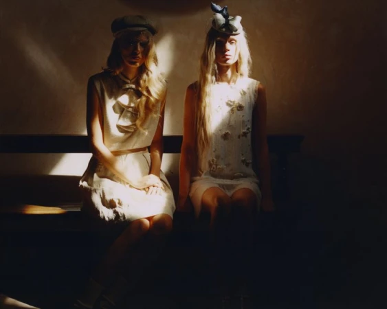 Two women in cream sleeveless dresses and vintage hats sit on a wooden bench illuminated by a sharp beam of light.