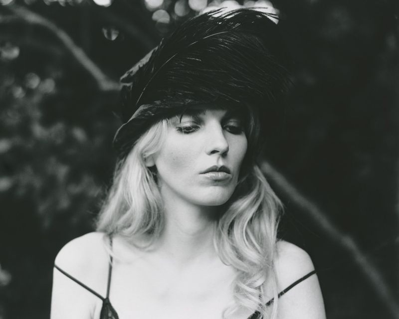 Black and white portrait of a blonde woman wearing a large black feathered hat and a strappy dark camisole top outdoors.