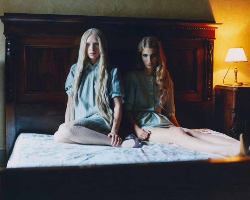 Two young adult females pose on a bed in front of a dark wood headboard with a lamp on a bedside table providing soft, warm light.