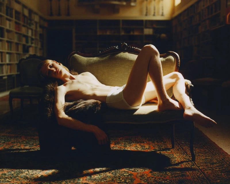 Indoor portrait of a shirtless young adult reclining on an antique couch in a library with bookshelves in the background.