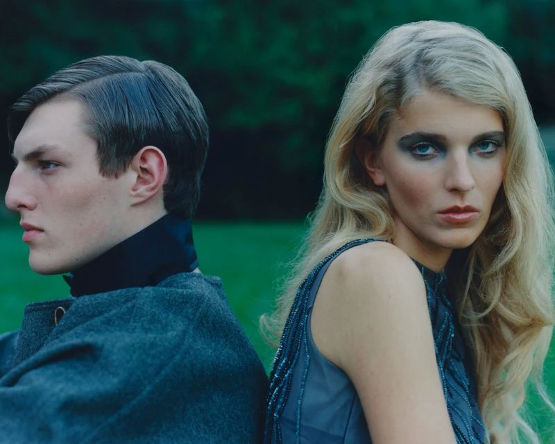 A fashion-style outdoor portrait of an adult male in a grey coat and an adult female in a grey beaded dress, back-to-back, looking at the camera.