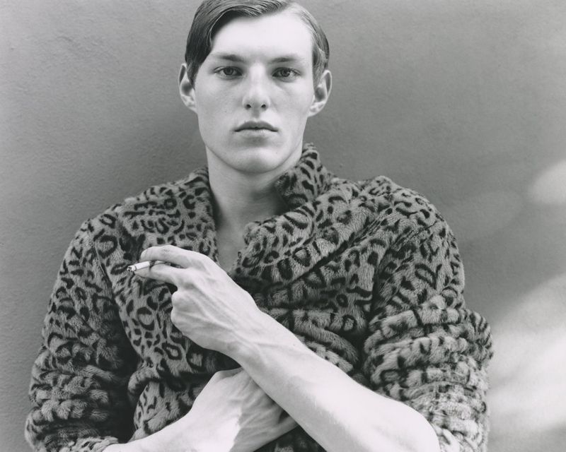 Monochrome portrait of a fair-skinned man wearing a leopard-print coat, holding a cigarette against a neutral gray background.