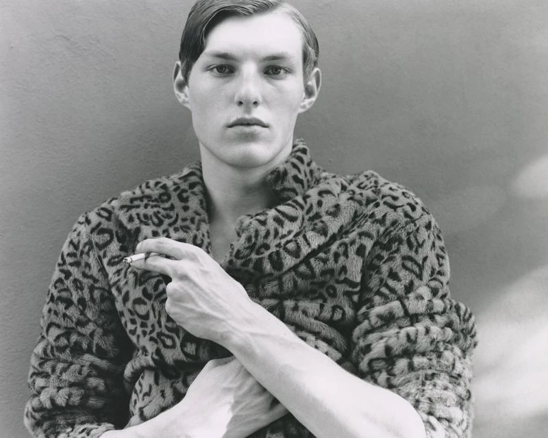 Monochrome portrait of a fair-skinned man wearing a leopard-print coat, holding a cigarette against a neutral gray background.