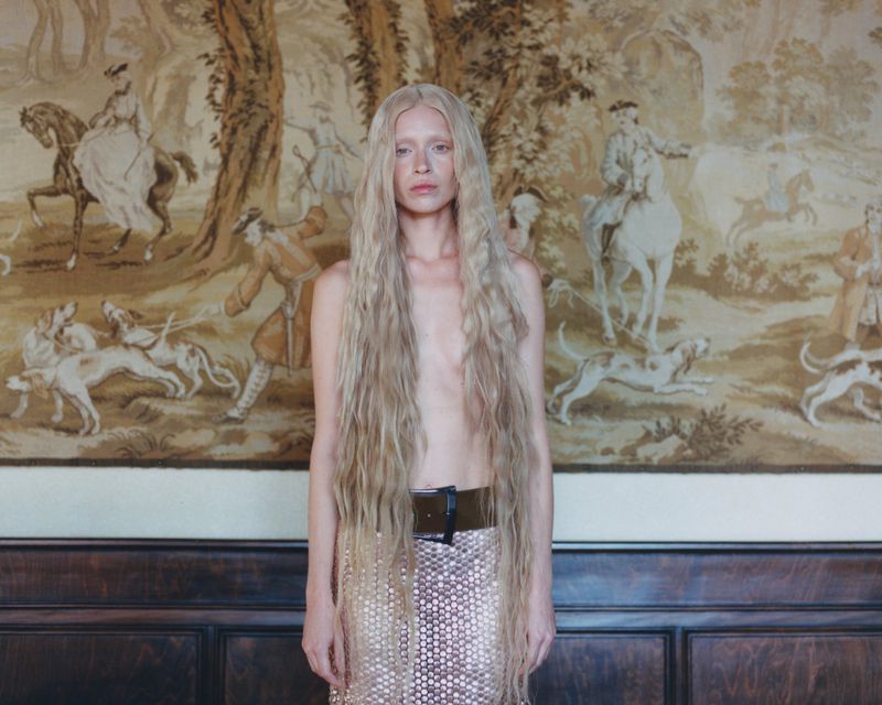 Adult female with long blonde hair poses in front of a tapestry depicting horses and hunting dogs, wearing a belt and beaded skirt.