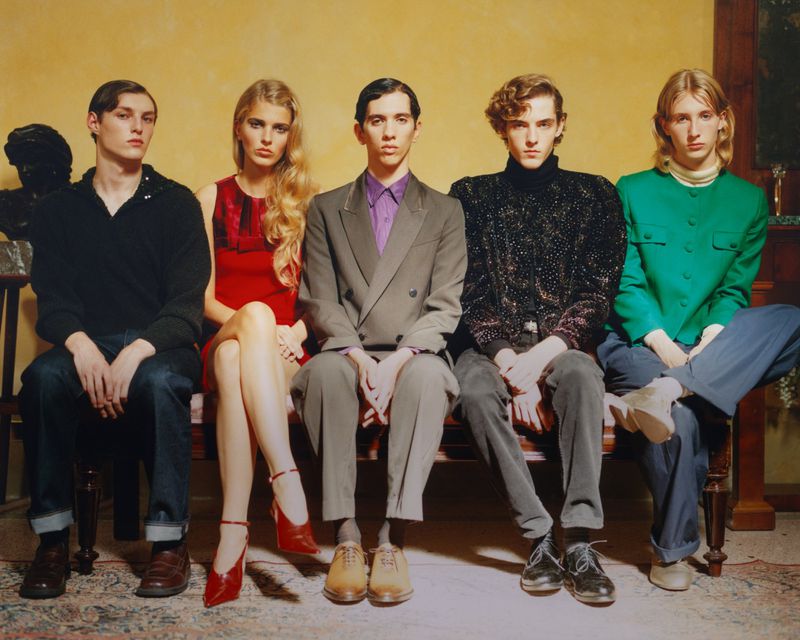 Group portrait of five fashionably dressed young adults sitting on a bench against a yellow wall with a statue in the background.