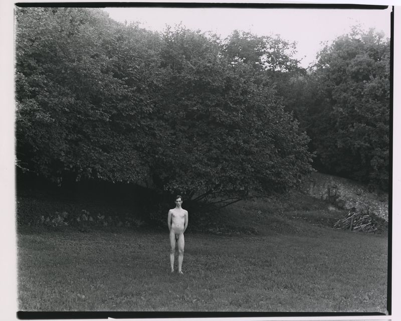 Full-length black and white photograph of a nude man standing in a grassy meadow in front of dense trees.