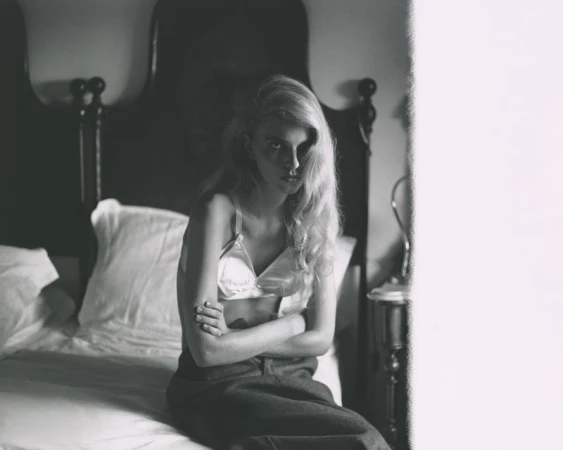 Monochrome photograph of a blonde woman sitting on a bed with folded arms, wearing a light satin bra and dark trousers.