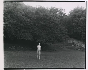 Full-length black and white photograph of a nude man standing in a grassy meadow in front of dense trees.