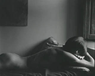 Black and white photograph of a woman with a dark bob haircut lying prone on a bed near a window with soft directional light.