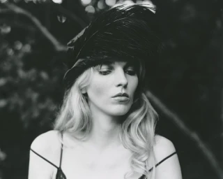 Black and white portrait of a blonde woman wearing a large black feathered hat and a strappy dark camisole top outdoors.