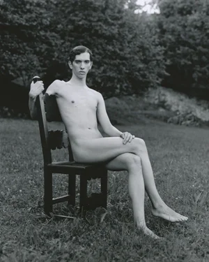 Black and white photograph of a nude man seated on a dark wooden chair on a grassy lawn with trees in the background.