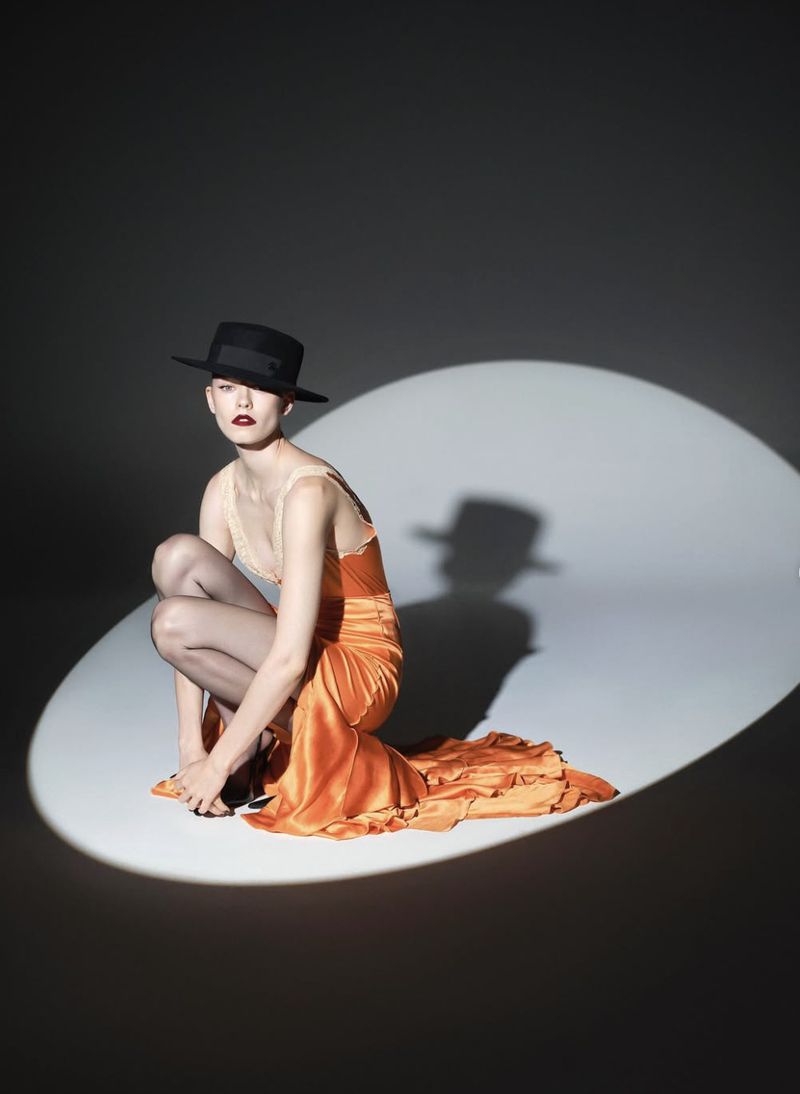 Woman in an orange dress and black hat is illuminated by a spotlight in a photo studio. Fashion shoot against a dark backdrop.