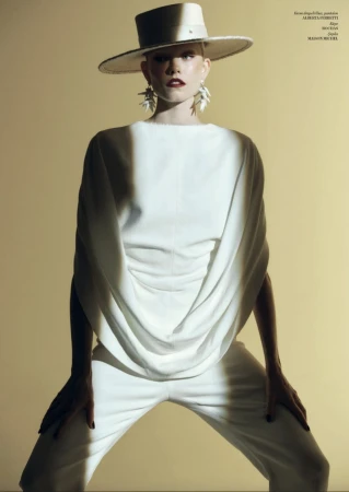 A bald woman in a white top and pants poses indoors, wearing a hat and statement earrings against a light yellow backdrop in a fashion shot.