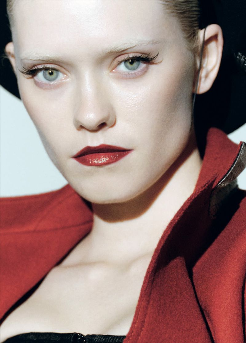 Fashion portrait of a woman with dramatic makeup, red coat, and black hat. The lighting is bright and the background is neutral.