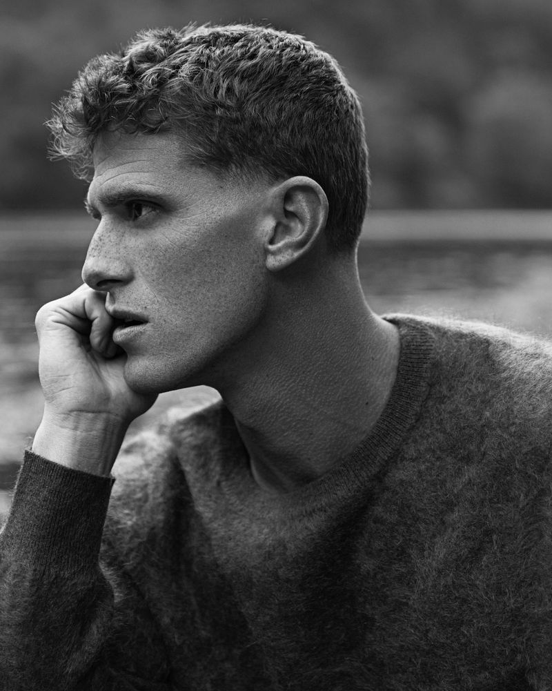Black and white portrait of a man with short curly hair wearing a sweater, resting his face on his hand in an outdoor environment.