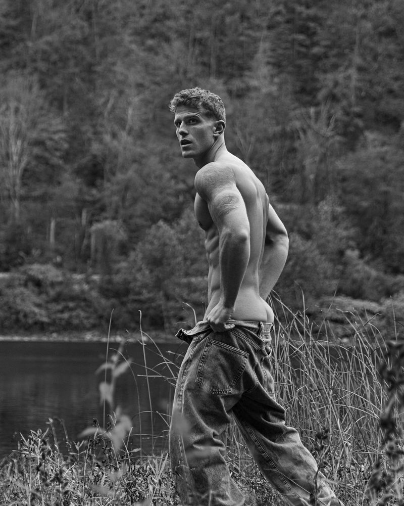Shirtless man wearing loose denim jeans standing outdoors near water and tall grass in black and white.