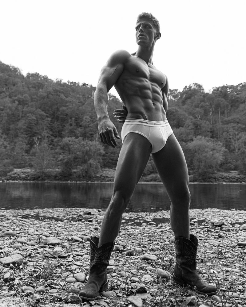 Black and white shot of a muscular man wearing briefs and cowboy boots standing on a rocky riverbank with a forested background.
