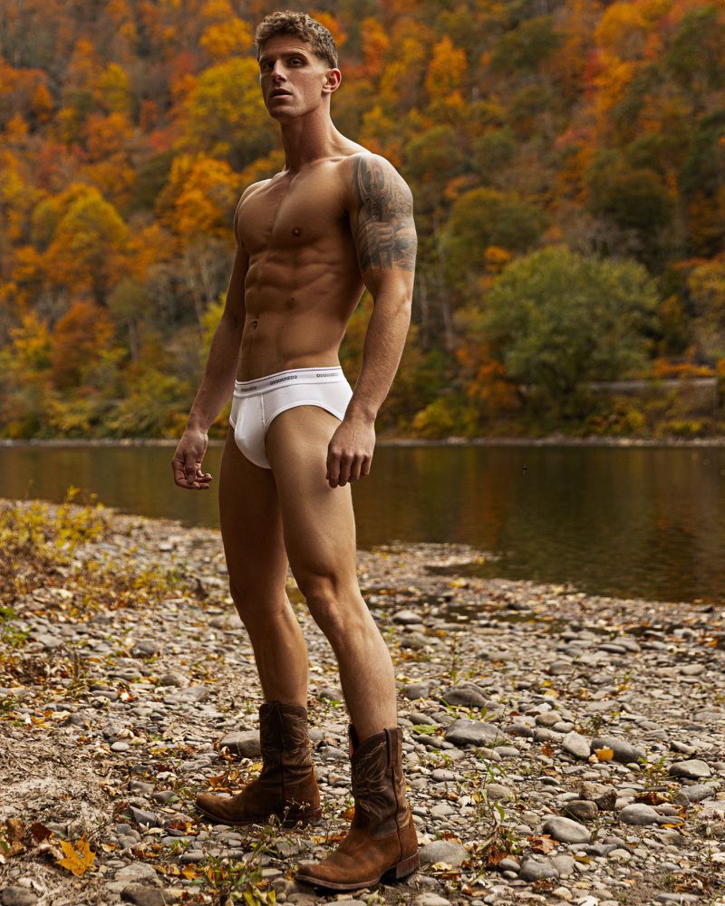 Athletic man poses shirtless in white briefs and cowboy boots on a rocky riverbank, surrounded by fall foliage and a calm river.