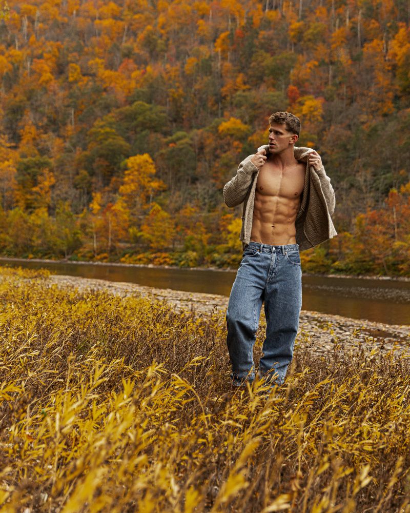 Male model poses outdoors near river in autumn. Shirtless man wears jeans and jacket in a scenic fall landscape with trees and foliage.