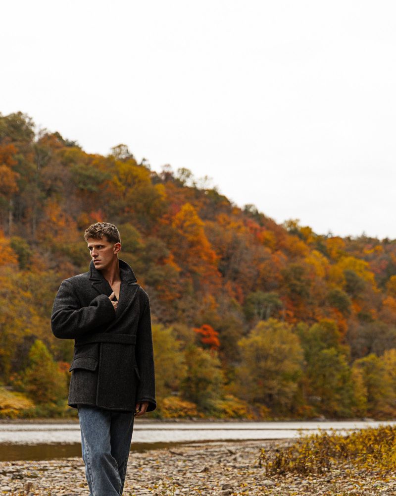 A young adult male model in a pea coat stands near a river with fall foliage backdrop, showcasing fashion and outdoor style in natural light.