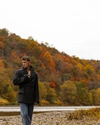 Model Matt van de Sande standing on a rocky riverbank in a dark belted coat and jeans with autumn foliage behind him.