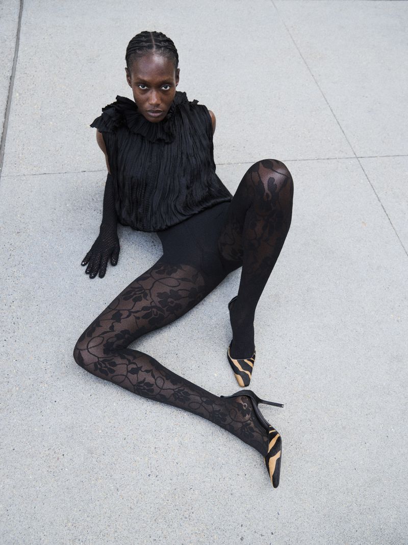 A fashionable Black woman poses, seated on grey concrete. She is wearing a black top, gloves, floral lace leggings, and zebra-print heels.