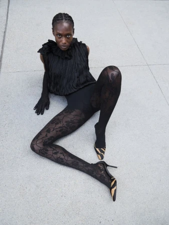 A fashionable Black woman poses, seated on grey concrete. She is wearing a black top, gloves, floral lace leggings, and zebra-print heels.