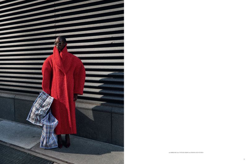 Woman in a red coat and sunglasses stands next to an urban building, holding a blue plaid bag for a fashion editorial photograph.