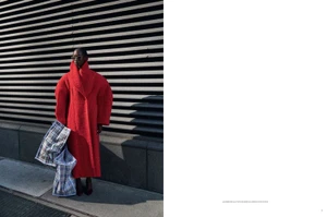 Essence Taylor wearing an oversized bright red coat and sunglasses, holding plaid tote bags in front of a slatted wall in hard sunlight.