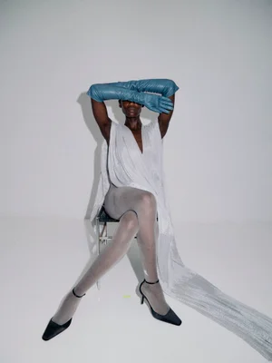 Model Essence Taylor seated in draped white fabric, long blue leather gloves over her eyes, sheer grey tights, and black heels.