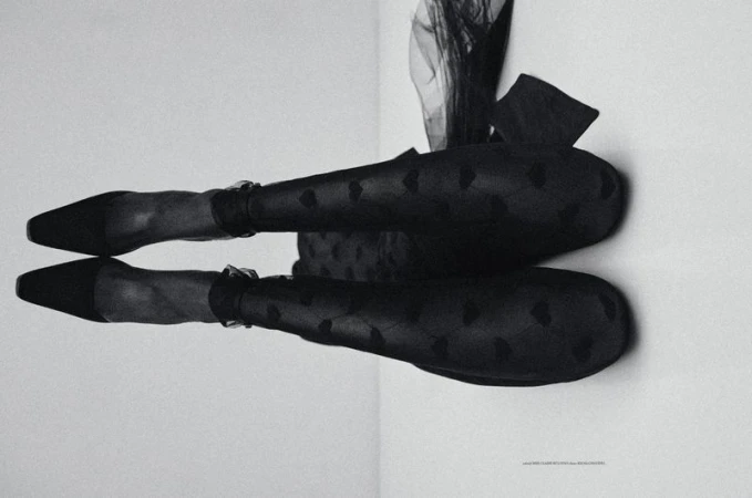 Close-up of a model's legs wearing sheer patterned black tights and pointed-toe black high heels against a pale background.