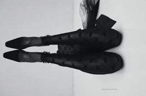 Close-up of a model's legs wearing sheer patterned black tights and pointed-toe black high heels against a pale background.