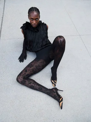 Model Essence Taylor sits on a concrete floor wearing a black ruffled top, sheer floral lace tights, and animal-print pointed pumps.