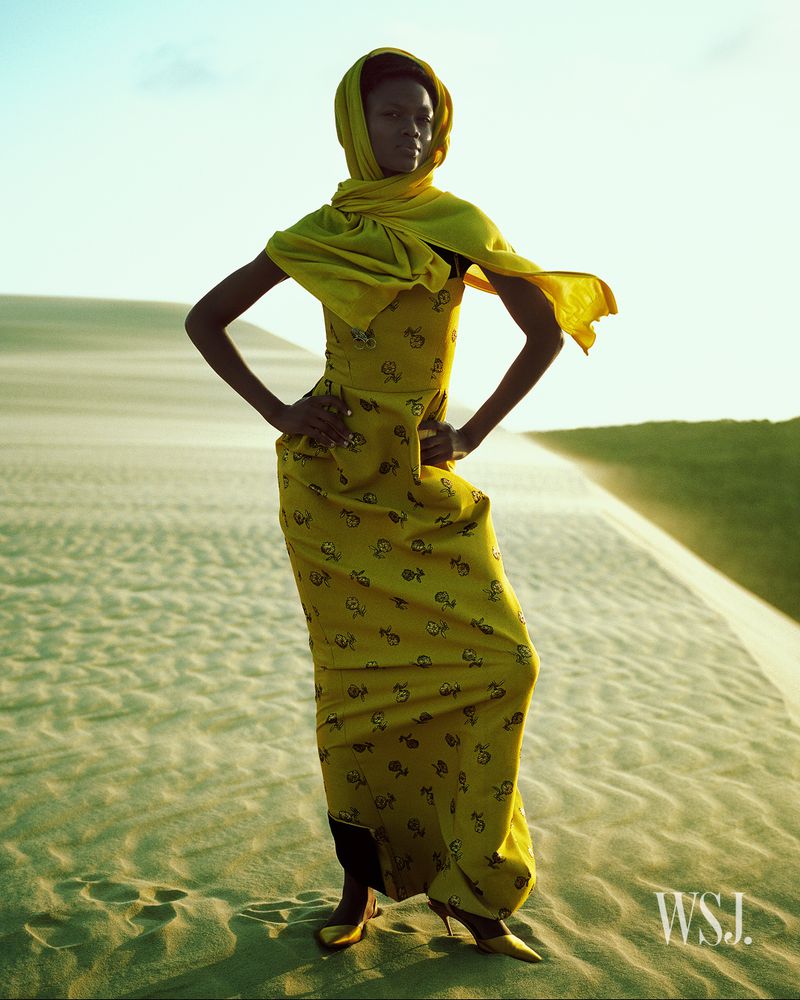 Fashionable woman in a yellow dress and headscarf poses on a sandy dune under a clear sky. The light creates a warm, serene atmosphere.