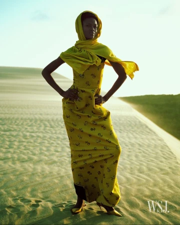 Fashionable woman in a yellow dress and headscarf poses on a sandy dune under a clear sky. The light creates a warm, serene atmosphere.