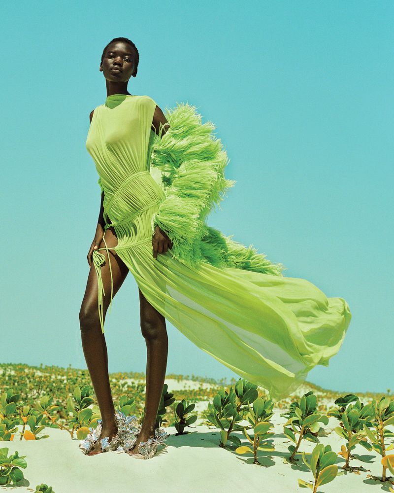 Model in flowing lime green feathered dress standing on sandy ground against a clear blue sky