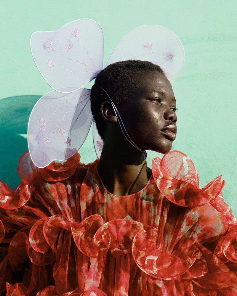 Black model in a voluminous red ruffled organza dress wearing sheer butterfly wings against a turquoise background.