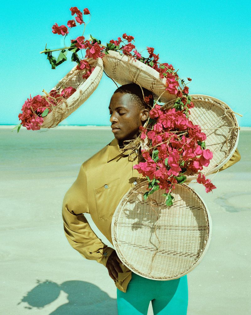 Portrait of a dark-skinned man on a beach, wearing baskets of bright pink flowers, a mustard jacket, and aqua leggings against a clear sky.
