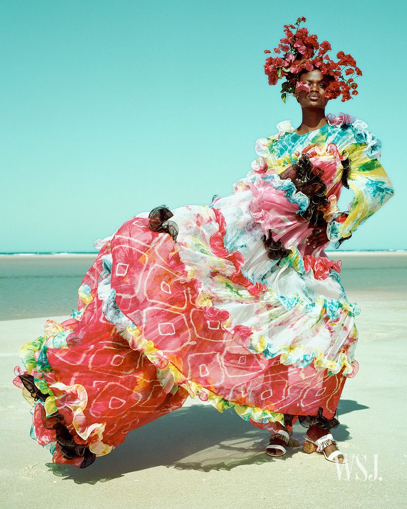 A full-length portrait of a woman on a beach wearing a vibrant, floral headpiece and a ruffled, patterned dress, posing in bright, sunny conditions.