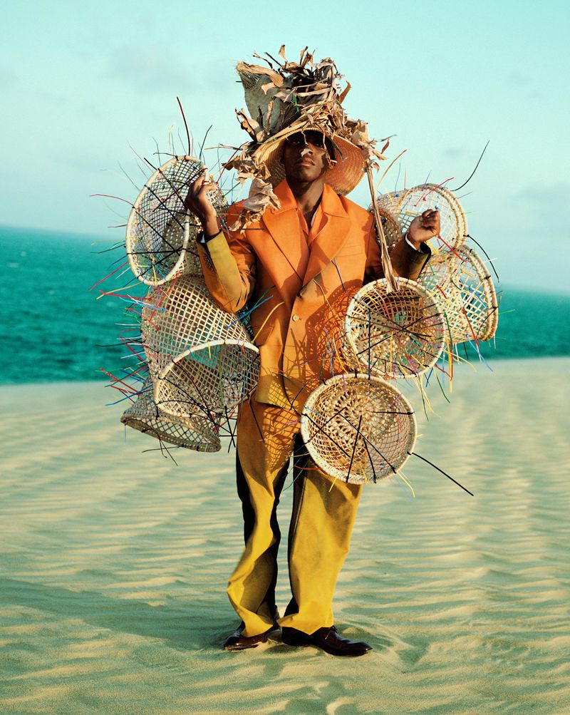 A man in an orange suit and elaborate woven headdress stands on a sand dune near the ocean wearing woven baskets as adornment.