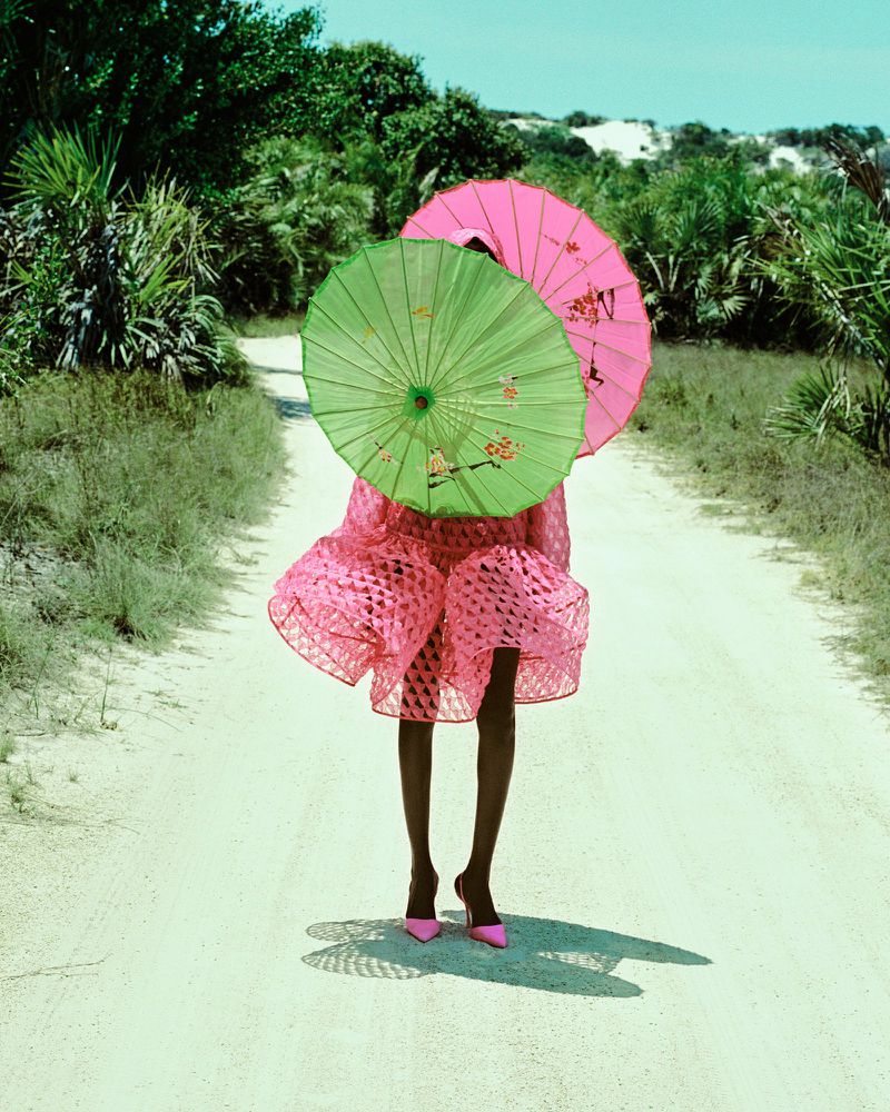 Woman in pink dress stands on sandy path, face obscured by green and pink parasols, set against trees and a blue sky.