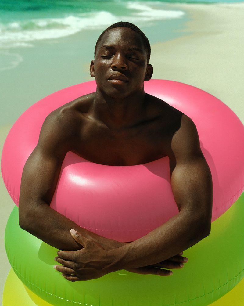 An adult Black male with eyes closed poses on a sandy beach, surrounded by colorful inflatable swim rings on a sunny day.
