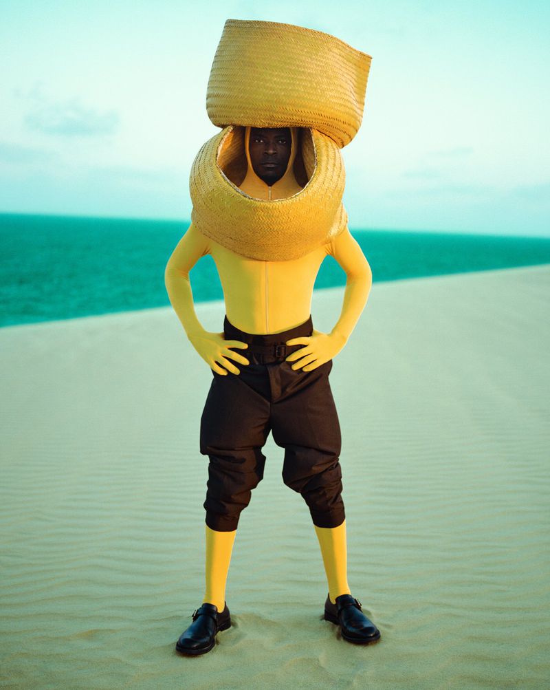 Person in a yellow jumpsuit wearing straw hats in a surreal fashion pose on a sand dune with sea and pale sky in the background.