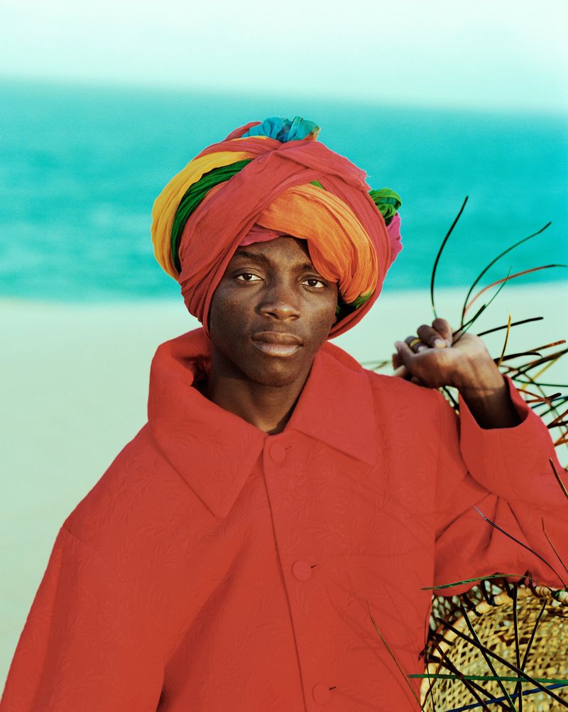 A dark-skinned person wearing a colorful turban and red coat poses on a beach, with a serene turquoise sea in the background.
