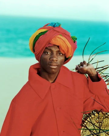 A dark-skinned person wearing a colorful turban and red coat poses on a beach, with a serene turquoise sea in the background.