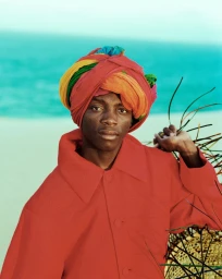 Portrait of a black man wearing a vibrant multicoloured turban and red jacket against an ocean backdrop